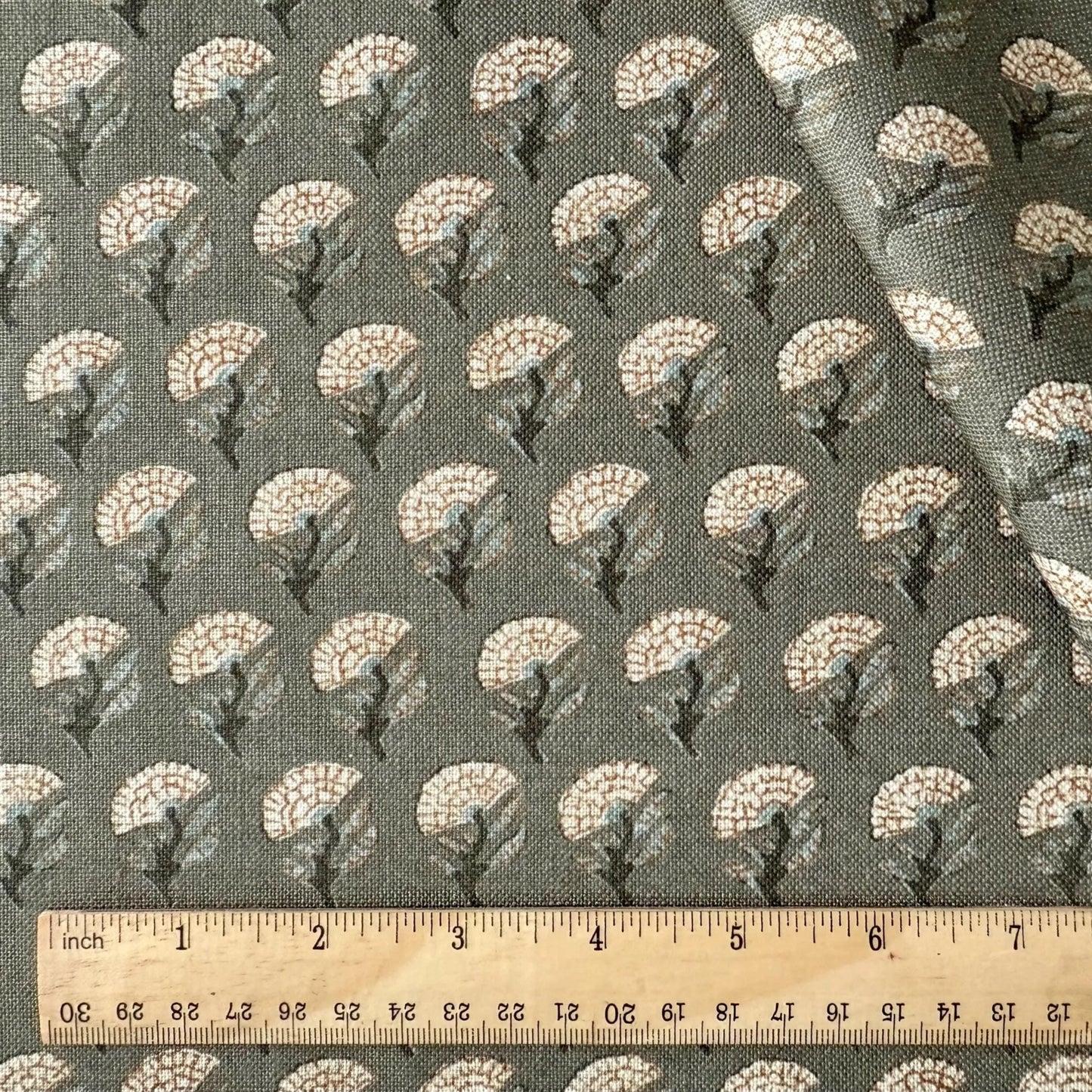 a fabric with a repeating pattern of small white flowers or leaves on a dark background. The fabric is laid out on a wooden ruler for size comparison.