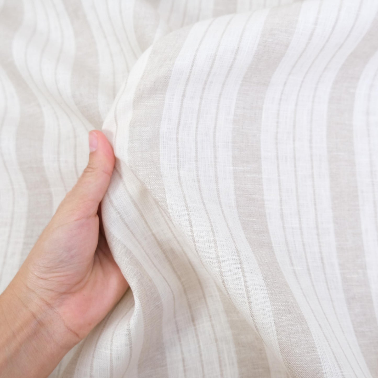 Hand holding taupe striped linen fabric to show scale, texture, and drape. This fabric is suitable for rustic farmhouse interiors, cottage style curtains, and neutral table linens. It is a breathable, natural-texture fabric with versatile taupe and white stripes, making it perfect for sewing projects such as pillow covers, kitchen textiles, DIY home décor, and lightweight craft projects with a soft yet durable finish.