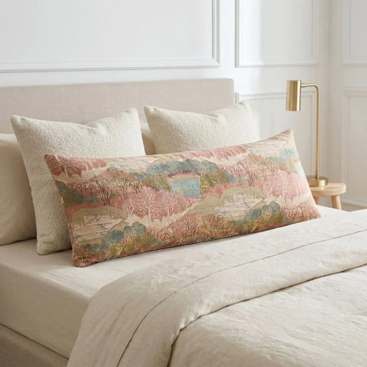 a bed with a large, colorful pillow featuring a landscape design. The pillow is placed on a white bedspread, and there is a lamp on a side table next to the bed.