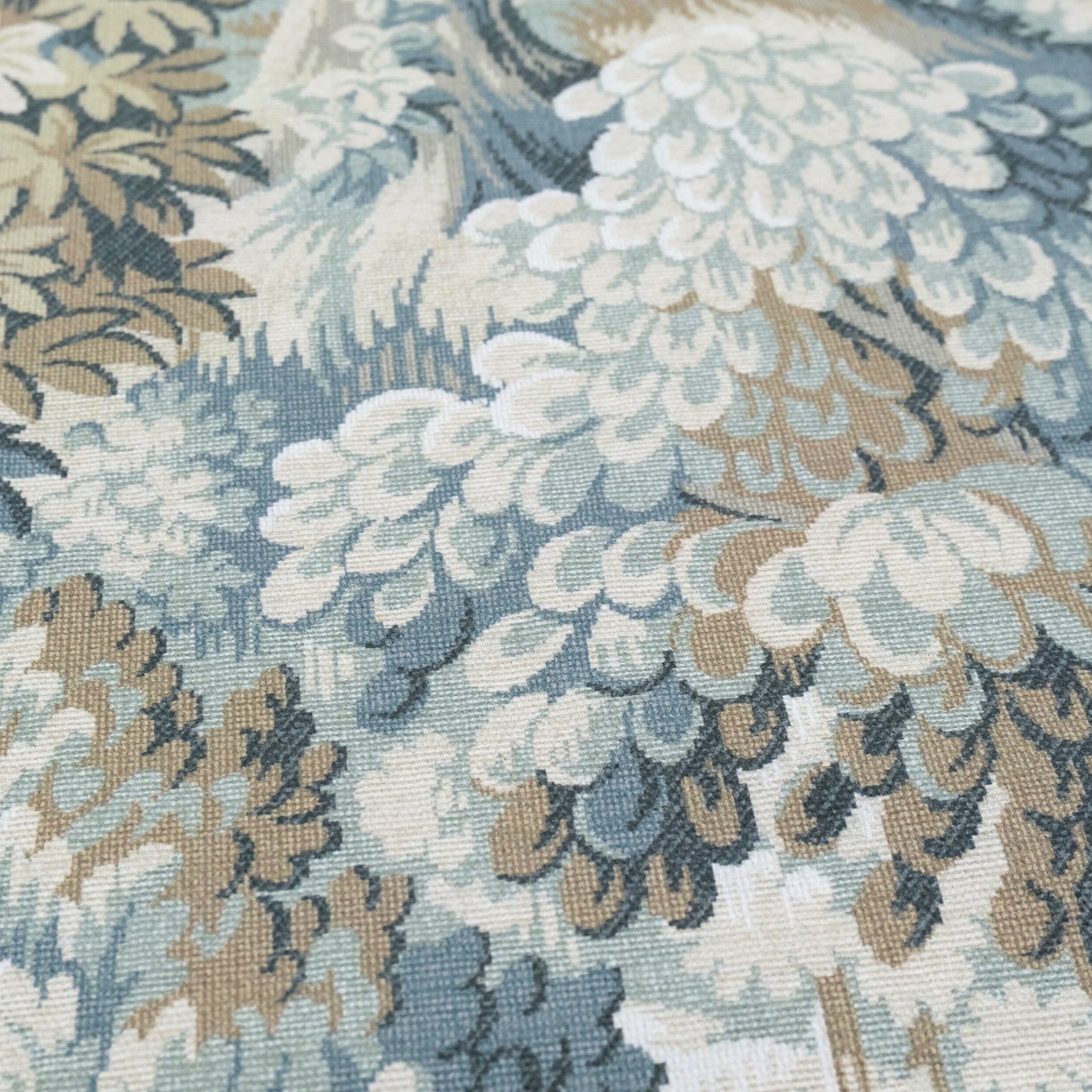 The image is a close-up of a fabric with a floral pattern in shades of blue, green, and brown. The pattern features large, intricate white and green feathers or leaves, creating a visually striking and detailed design.