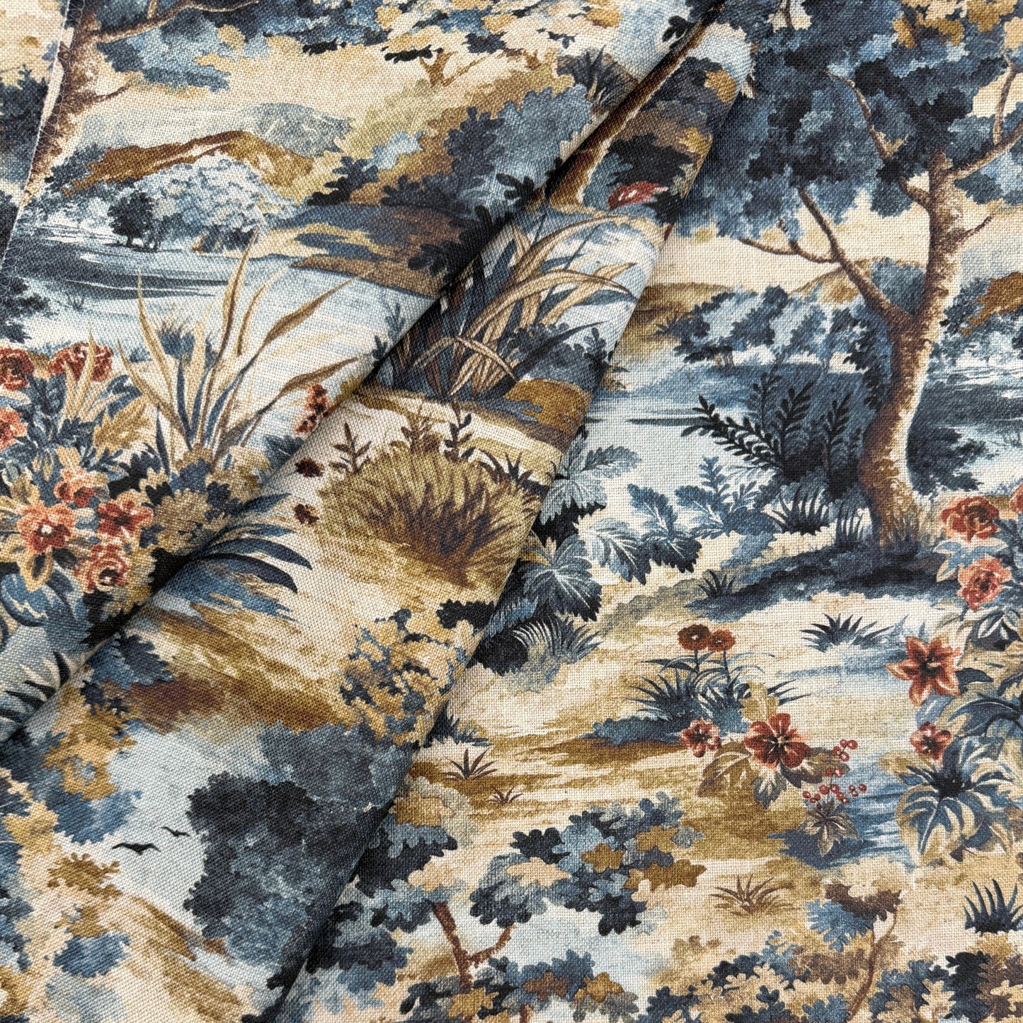 The image is a close-up of a fabric with a detailed floral and landscape pattern. The fabric features a variety of colors, including shades of blue, green, and brown, as well as various types of flowers and foliage. The pattern appears to be a repeating design, with the elements of the landscape and floral motifs interspersed throughout the fabric.