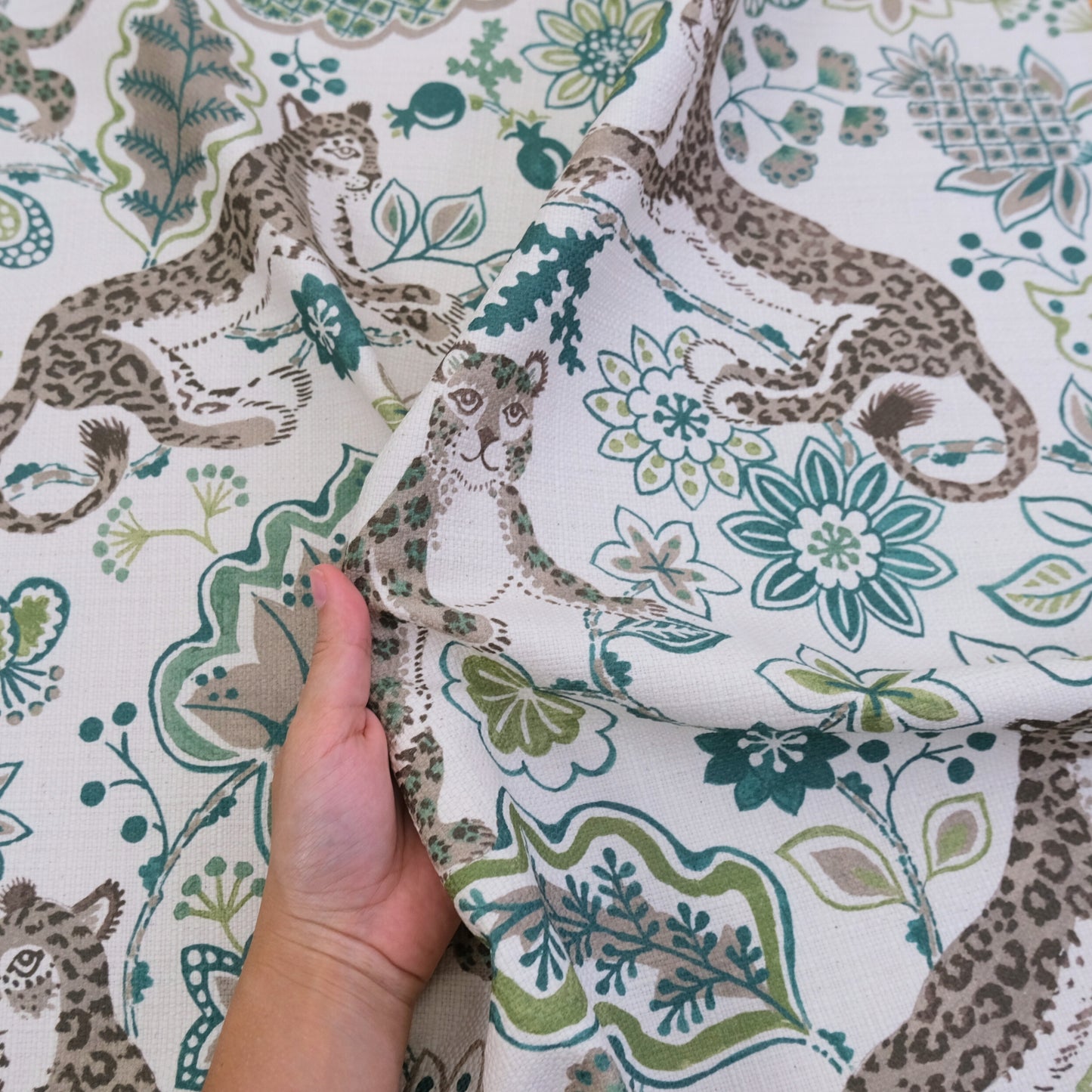 A hand is holding up a fabric with a repeating pattern of leopard and floral designs in shades of green and gray.