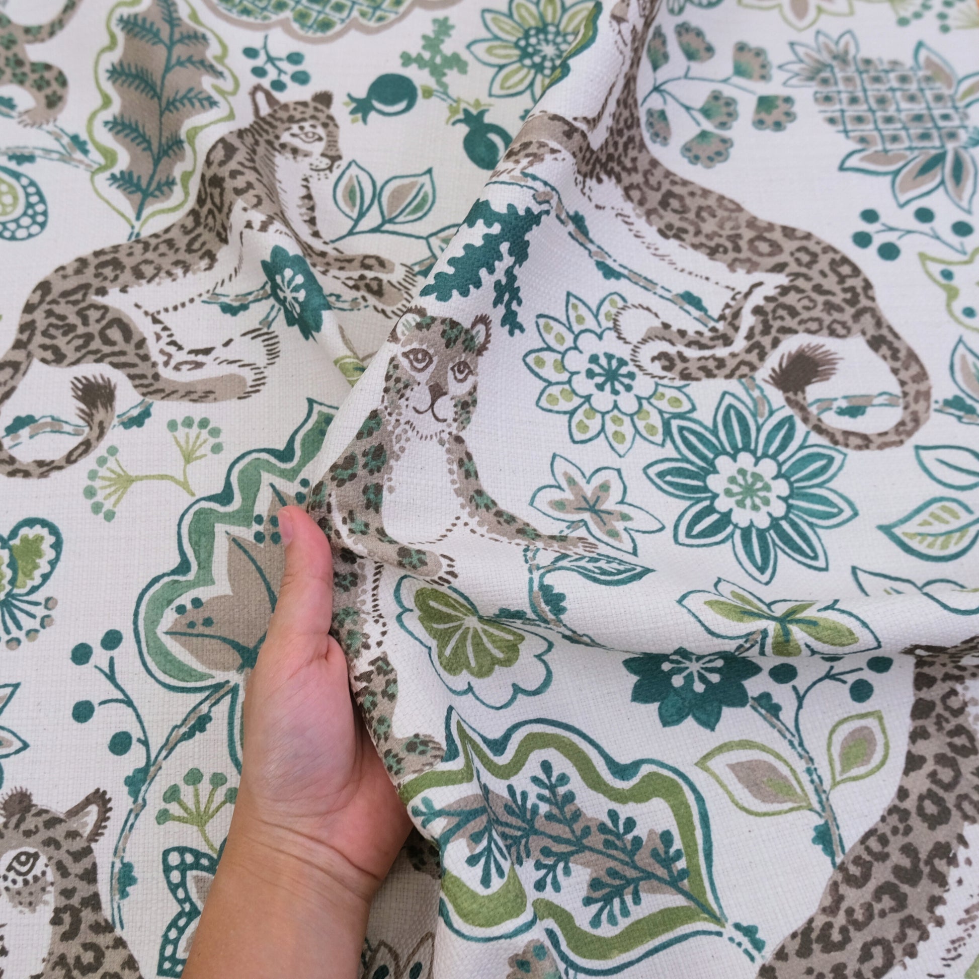 A hand is holding up a fabric with a repeating pattern of leopard and floral designs in shades of green and gray.