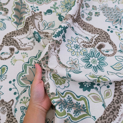 A hand is holding up a fabric with a repeating pattern of leopard and floral designs in shades of green and gray.