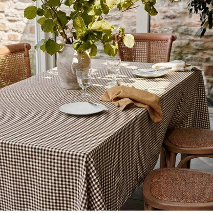 a table set for a meal, with a checkered tablecloth, plates, glasses, and a vase of leaves. The table is surrounded by chairs, creating a cozy dining atmosphere.